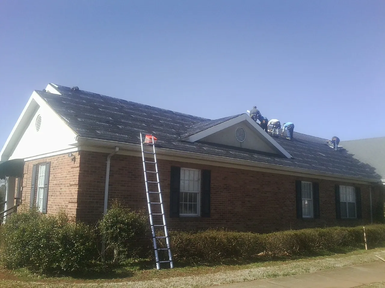 Crew tearing shingles off a brick ranch roof with a ladder set — Emergency Roof Repair work by our crew in Colmar