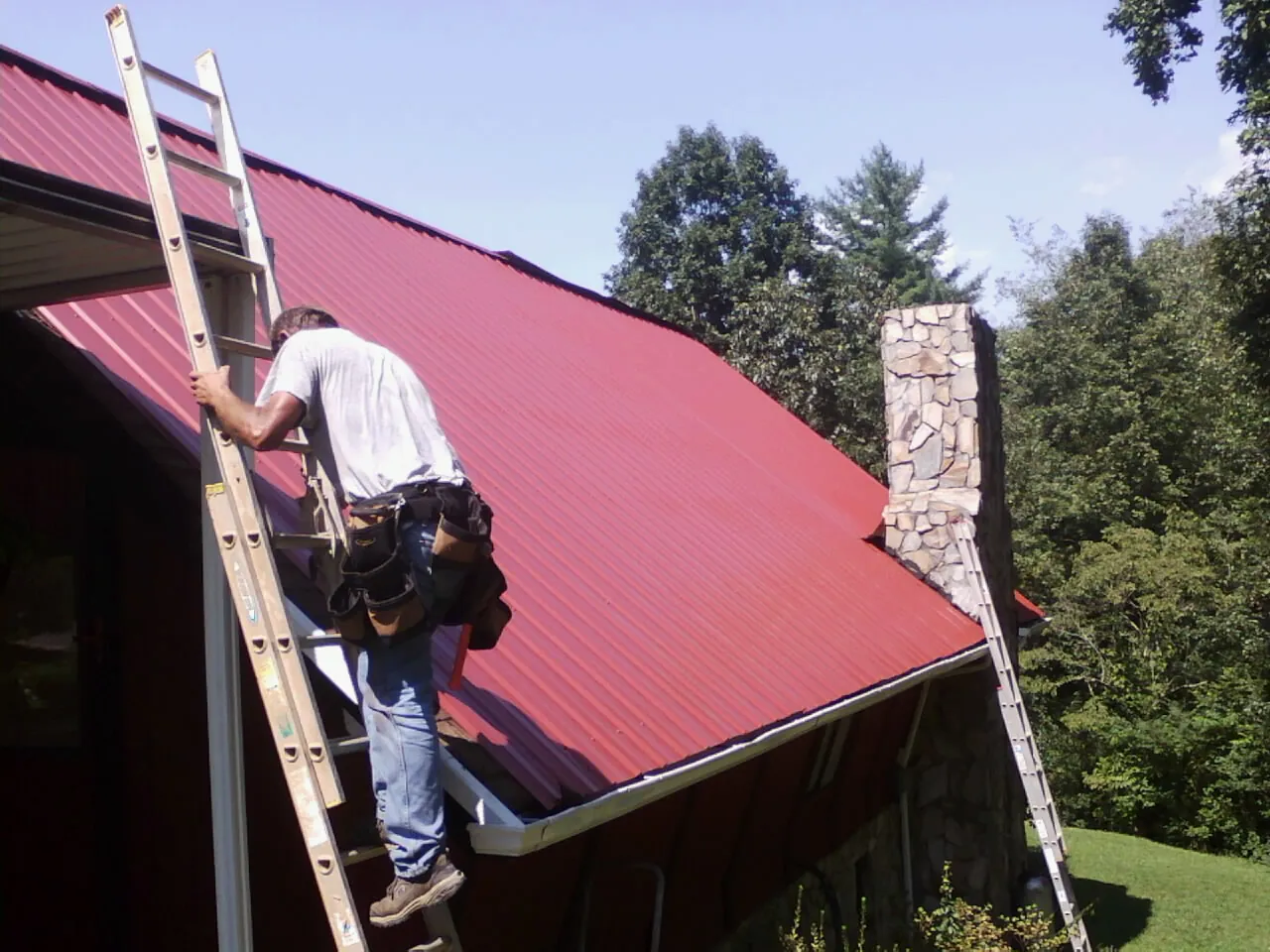 Roofer on a ladder beside a red metal roof and stone chimney — Soffit & Fascia Repair by Roofing Brothers in Colmar
